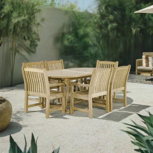 70002 Montserrat 7 piece teak oval Dining Set of Monsterrat Oval Teak Dining Table and 6 Veranda Dining Chairs angled on rock patio surrounded by plants with privacy wall in background