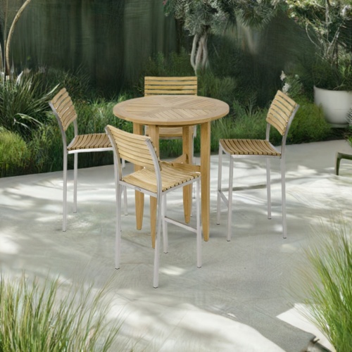 70683 Laguna Round Vogue 5 pc Teak Barstool Set of Laguna 36 inch round teak bar table and 4 Vogue Side Bar Stools angled on concrete patio surrounded by plants with fence in background