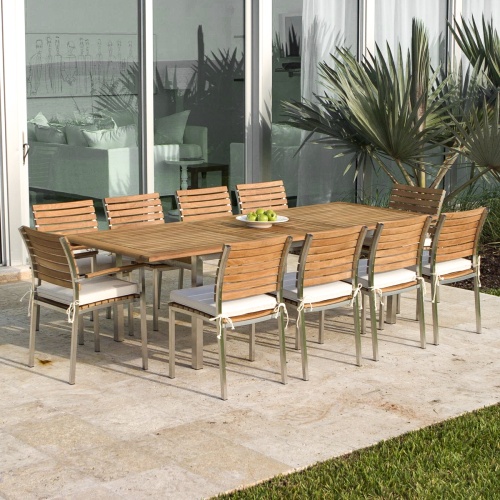 70937 Vogue stainless steel and teak rectangular dining set of Vogue Rectangular Dining Table and 2 Dining Arm Chairs and 8 Side Chairs with optional seat cushions angled on patio with home and plants in background