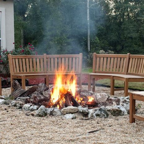 13852 Buckingham 6 foot Teak Bench showing two around a campfire with plants & trees in background 