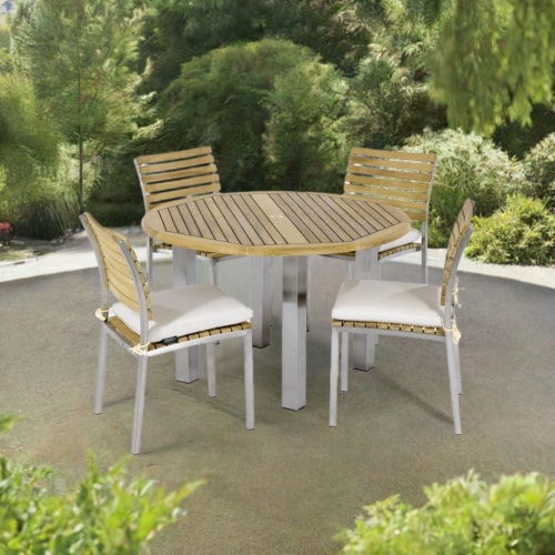 70438 Vogue Dining Set for 4 of Vogue Teak and Stainless Steel 48 inch Round Dining Table and 4 Stacking Dining Chair with optional seat cushions angled on patio surrounded by plants and trees