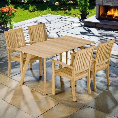 70564 Surf Sussex 5 pc Dining Set of Surf 5ft Rectangular Table and 4 Sussex Dining Chairs angled on stone patio with landscape plants and grass and a lit outdoor fireplace in the background