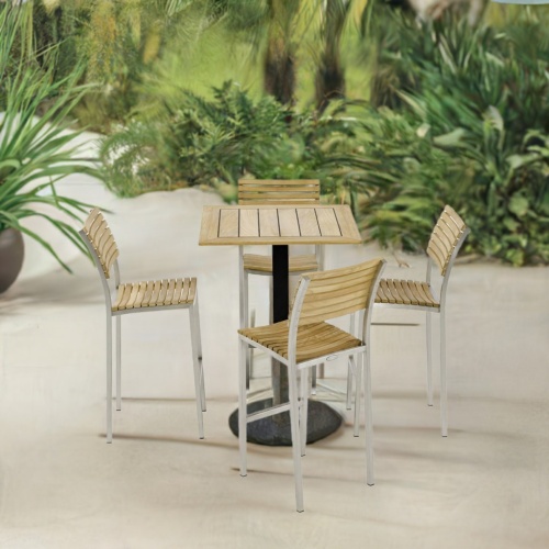 70687 Vogue 5 pc Bar Furniture Set of a Teak 30 inch Square Table Top on black base and 4 Vogue Teak and Stainless Steel Barstools aerial side view on patio with plants in background