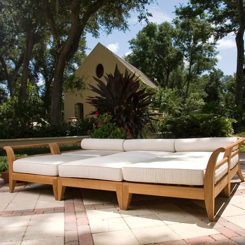 70100 Aman Dais 6 piece teak daybed set with cushions front angled showing two corner three end pieces and one ottoman on stone patio with trees plants and house in background