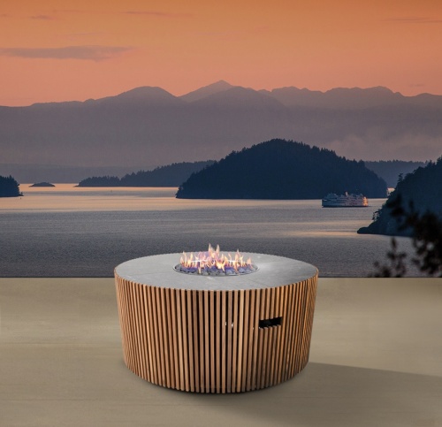 14100 Agora round Fire Table lit in side angled view on outdoor terrace overlooking a boat on a lake with mountains and islands and sunset view in background 