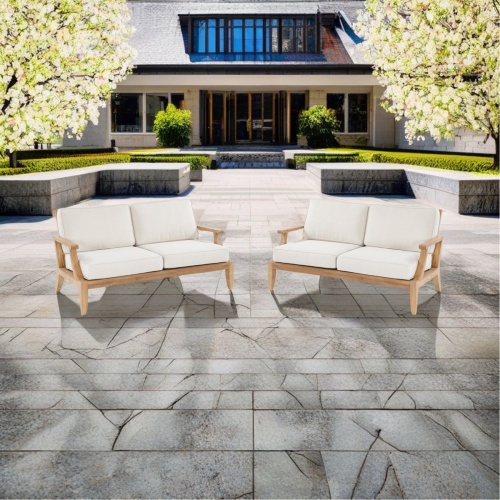 70914 two laguna teak loveseats with cushions angled front view on stone patio with flowering trees and house in the background 