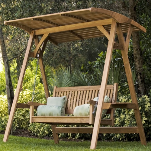 13955bo veranda teak swinging bench with teak canopy with four toss pillows with landscape plants and trees in background