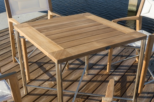 25815 Odyssey Teak and Stainless Steel 32 Inch Square Outdoor Folding Table and 2 Odyssey Chairs closeup angled view on sandy beach with large rocks in background