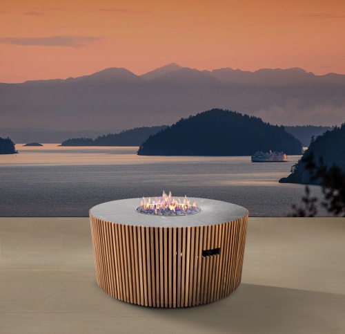 14100 Agora round Fire Table lit in side angled view on outdoor terrace overlooking a boat on a lake with mountains and islands and sunset view in background